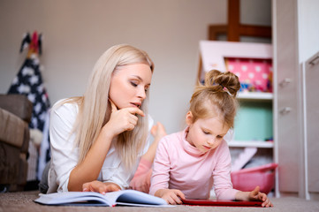 Young mother reads a book at home lying on the floor at home with her daughter, the child watches cartoons on the tablet and plays, happy family, love, family concept.