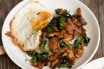 Pad Kra Pao Beef, (Stir fried beef with thai basil and chilli) top up with sun rise side up eggs on the white dish. close up , wooden table