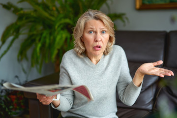 Woman reading newspaper at home, shocked and surprised over the news. Mature woman.