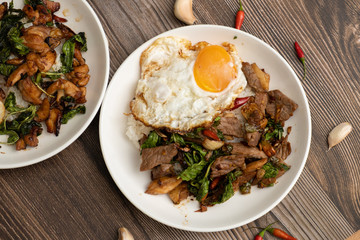 Pad Kra Pao Beef, (Stir fried beef with thai basil and chilli) top up with sun rise side up eggs on the white dish. close up , wooden table