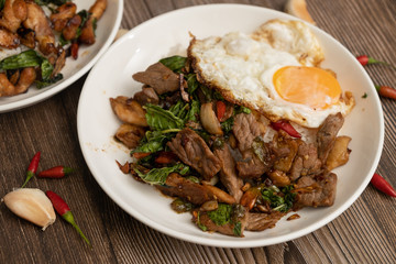 Pad Kra Pao Beef, (Stir fried beef with thai basil and chilli) top up with sun rise side up eggs on the white dish. close up , wooden table