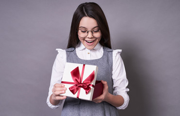 Beautiful young lady with long dark hair in glasses holds a box with gifts, on a gray background.