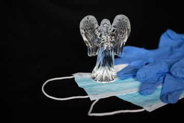 The crystal angel figurine stands on protective medical masks and latex blue gloves on a black...
