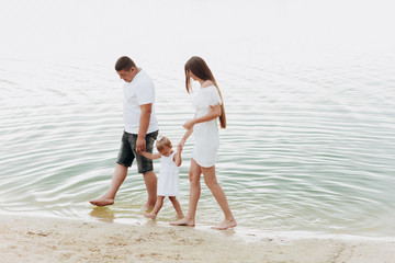 Mom, dad hugging daughter walking on the beach near lake. The concept of summer holiday. Mother's, father's, baby's day. Family spending time together on nature. Family look. Sun light.
