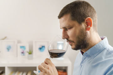 Side view portrait of adult caucasian man wearing shirt holding glass of red wine tasting at home at evening or night close up male with beard and short hair at home alone one