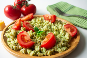 Basil pesto fusili pasta with Parmesan cheese and sliced tomatoes in a wooden plate and a green napkin with a fork in the background