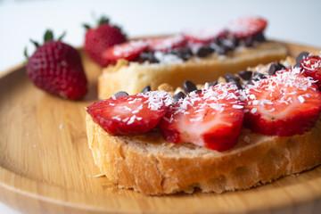 Peanut butter spread toasts with chocolate chips, strawberries, coconut flakes and homemade bread on a wooden plate