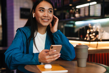 Photo of asian woman using mobile phone and wireless earphones in cafe