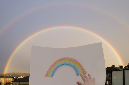 double rainbow seen from a window with a rainbow painted by a child showing his hand. The rainbow symbolizes support and hope to pass the COVID-19 epidemic