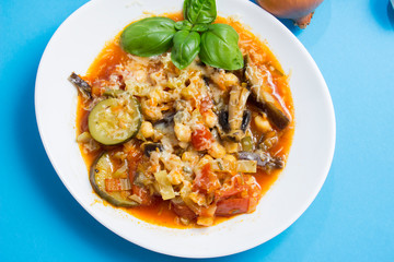 Vegetable stew with eggplant, zucchini, tomatoes and mushrooms. White plate and blue background