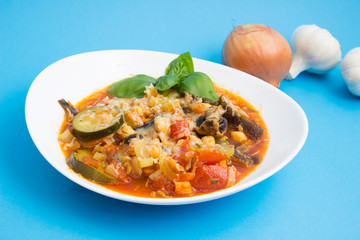 Vegetable stew with eggplant, zucchini, tomatoes and mushrooms. White plate and blue background