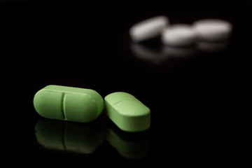 Green medical pills lie on a black table with a reflective surface. In the background are white pills.