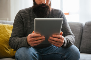 Young millennial man with beard checks the notifications of his tablet sitting on the sofa at home - Hipster is watching online videos on his device in a moment of relax