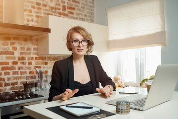 A young woman in glasses works remotely on a laptop in her kitchen. A blond girl gesticulating...