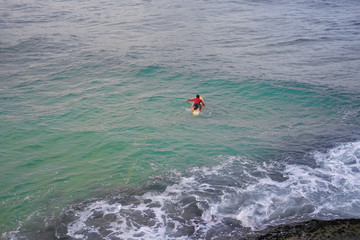 Surfer man in a red jacket with a surfboard in the azure ocean.