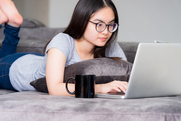Portrait beautiful Asian young woman lying and relax work with a laptop, Cute girl teens looking at the monitor intently on the sofa working remotely with an internet computer communication from home