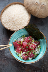 Ahi poke served in a turquoise bowl, vertical shot on a grey stone surface with raw rice in a coconut shell in the background