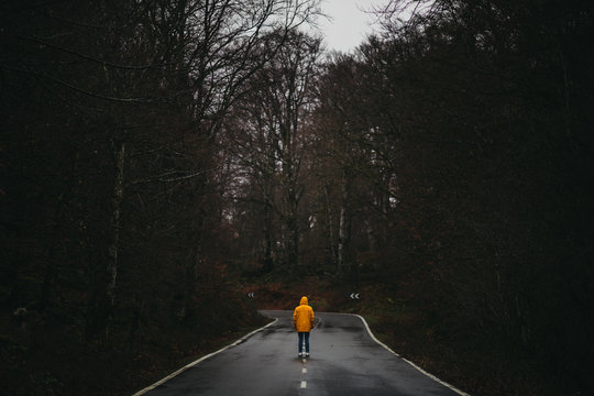 Back view of anonymous man in yellow jacket walking on empty asphalt road among green forest