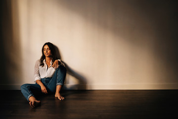 Gorgeous barefoot brunette in white shirt and blue jeans smiling with closed eyes while sitting on floor near wall in empty room