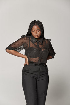 Attractive African American female in trendy transparent blouse and black trousers with closed eyes against gray background in studio
