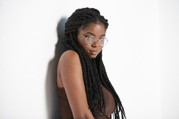 Young African American female with braids in stylish round glasses leaning on white wall and looking at camera