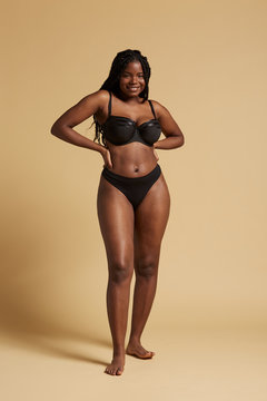 Curvy beautiful African American woman with braids in lingerie keeping hands on waist and looking at camera against yellow background