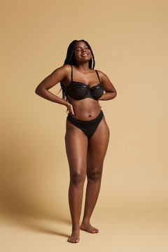 Curvy beautiful African American woman with braids in lingerie keeping hands on waist and looking at camera against yellow background