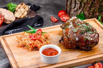 Pork knuckle on light wooden board served with stewed cabbage and barbecue sauce on dark concrete background