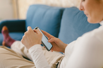 Faceless copped woman without shoes chilling on cozy sofa in office enjoying cup of coffee and surfing mobile phone