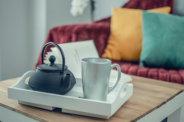 Closeup and selective focus on a tray with a teapot and a cup on a wooden table.  Burgundy blanket, yellow and green cushions on a grey sofa in the blurred background.