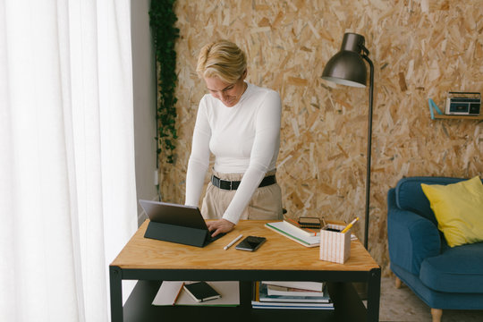 Casual modern blond businesswoman at wooden desk using tablet with keypad working in calm cozy office