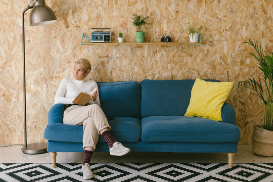Blonde woman with short hair in white shirt sitting on sofa looking away and writing in notebook working on business project
