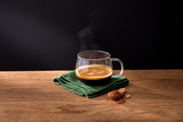Glass cup of aromatic hot black coffee on green napkin placed with spoon of ground cinnamon on wooden table with black background