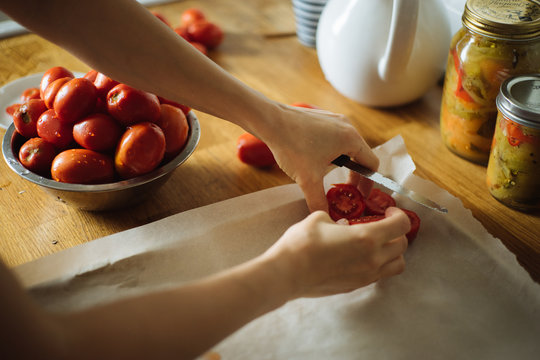 Crop housewife putting cut fresh tomatoes on baking tray while preparing traditional homemade preserves at wooden table in kitchen