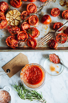 Top view of glass pot with homemade tomato sauce placed on table next to metal tray with grilled tomatoes with garlic and rosemary