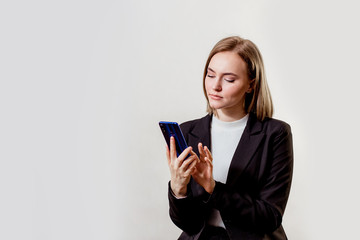 a young beautiful serious woman is sitting and writing something on the phone, dressed in a black jacket, on a gray background