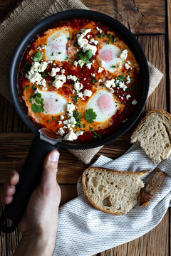 Top view of crop person putting on wooden table frying pan with delicious shakshuka made with fried eggs with vegetables and feta cheese