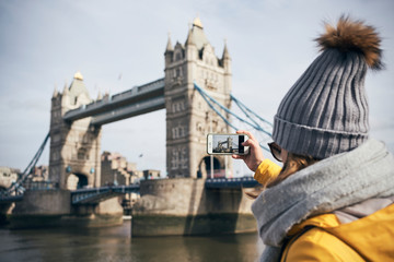 Back view of female traveler in warm clothes and hat taking picture of Tower bridge with smartphone during sightseeing in London in sunny day