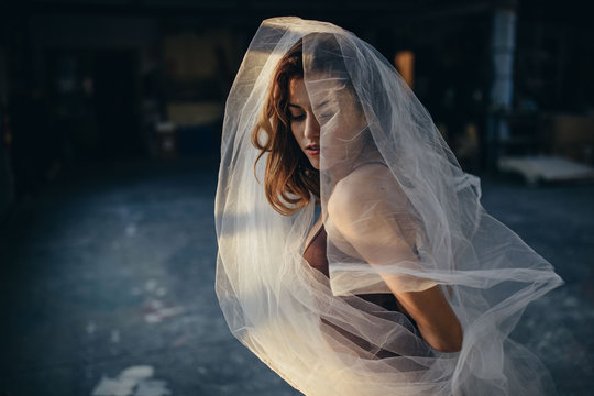 Young talented female dancer with closed eyes in bodysuit dancing with transparent veil while training alone in light studio