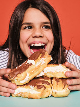Closeup of cheerful little girl in casual wear enjoying sweet eclairs with chocolate while sitting at table against red background