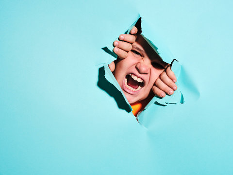 Screaming angry boy with grimace looking through ripped blue paper sheet in studio