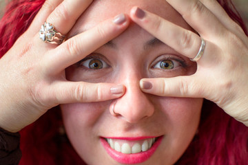 Close up portrait of a girl with green eyes and reddish hair covering her face with her hands revealing her eyes and her huge smile.