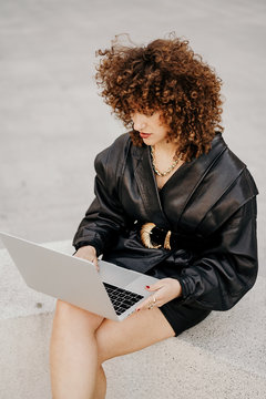 From above businesswoman in black leather outfit sitting on border and browsing data on laptop while working on remote project on city street