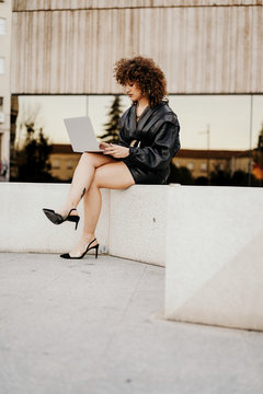 businesswoman in black leather outfit sitting on border and browsing data on laptop while working on remote project on city street