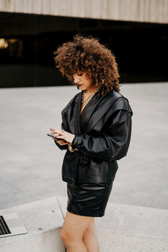 Side view of smart lady in vintage outfit browsing smartphone and standing near border with laptop while doing remote job on street