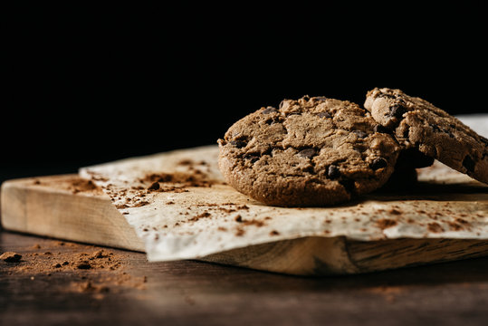 Delicious homemade crispy cookies with chocolate chips served on baking paper on wooden board against black background