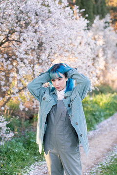 Young stylish female touching long blue hair looking at camera wearing trendy overall enjoying blooming tree while standing in spring garden