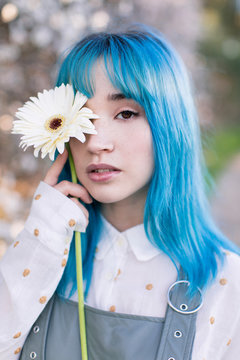 Modern trendy female with blue hair holding a fresh flower covering eye and looking at camera while standing in blooming spring garden