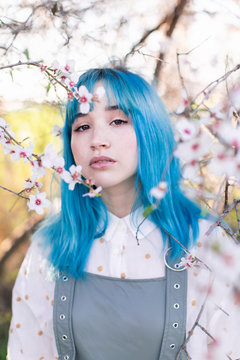 Young stylish female with long blue hair looking at camera wearing trendy overall enjoying blooming tree while standing in spring garden