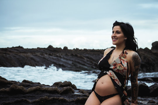 Sensual pregnant woman looking away wearing long waving lingerie robe sitting with bare belly on rocky coast in gloomy day